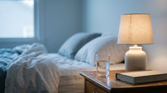 A glass of water on a nightstand at dusk beside a lamp and a book