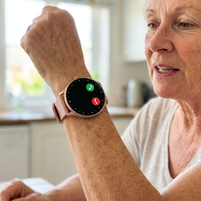 A woman in her sixties lifts her wrist to answer a call on the round 360 Health Tracker, its pink band and rose gold case catching the kitchen light, green and red call buttons on the screen